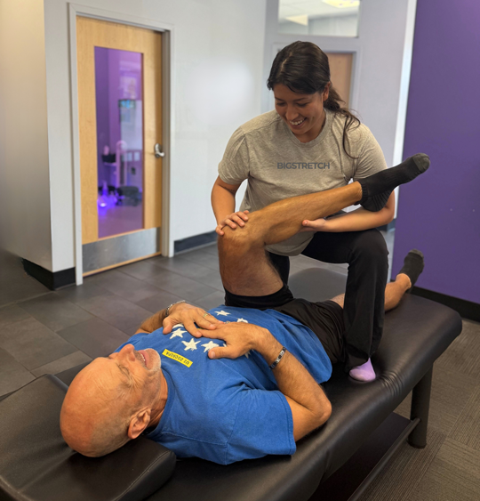 Trainer assisting an older man with a guided leg stretch during a one-on-one assisted stretch session at Bigstretch Encinitas, both focused and smiling in a professional studio setting.