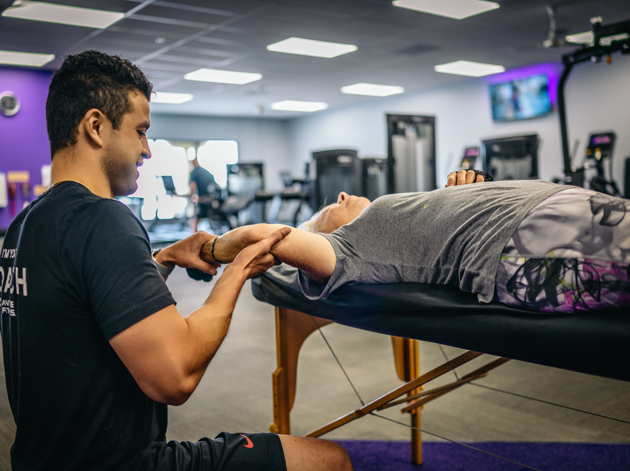 Assisted stretch therapy session at Bigstretch Encinitas, with a trainer guiding an older woman through a supported leg stretch on a therapy table in a gym setting