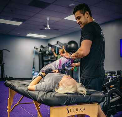 Assisted stretch therapy session at Bigstretch Encinitas, with a trainer guiding an older woman through a supported leg stretch on a therapy table in a gym setting.
